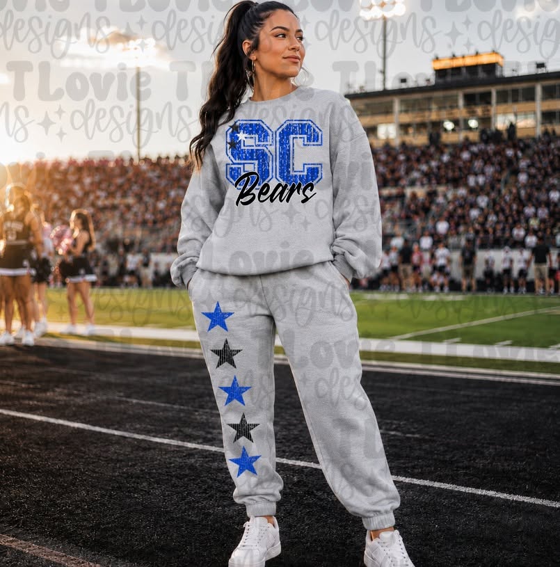 A woman stands on a football field wearing a light gray sweatshirt and jogger sweatpants set. The sweatshirt has a large blue and black 'SC Bears' graphic on the front, and the joggers have a vertical row of blue and black stars down the left leg.