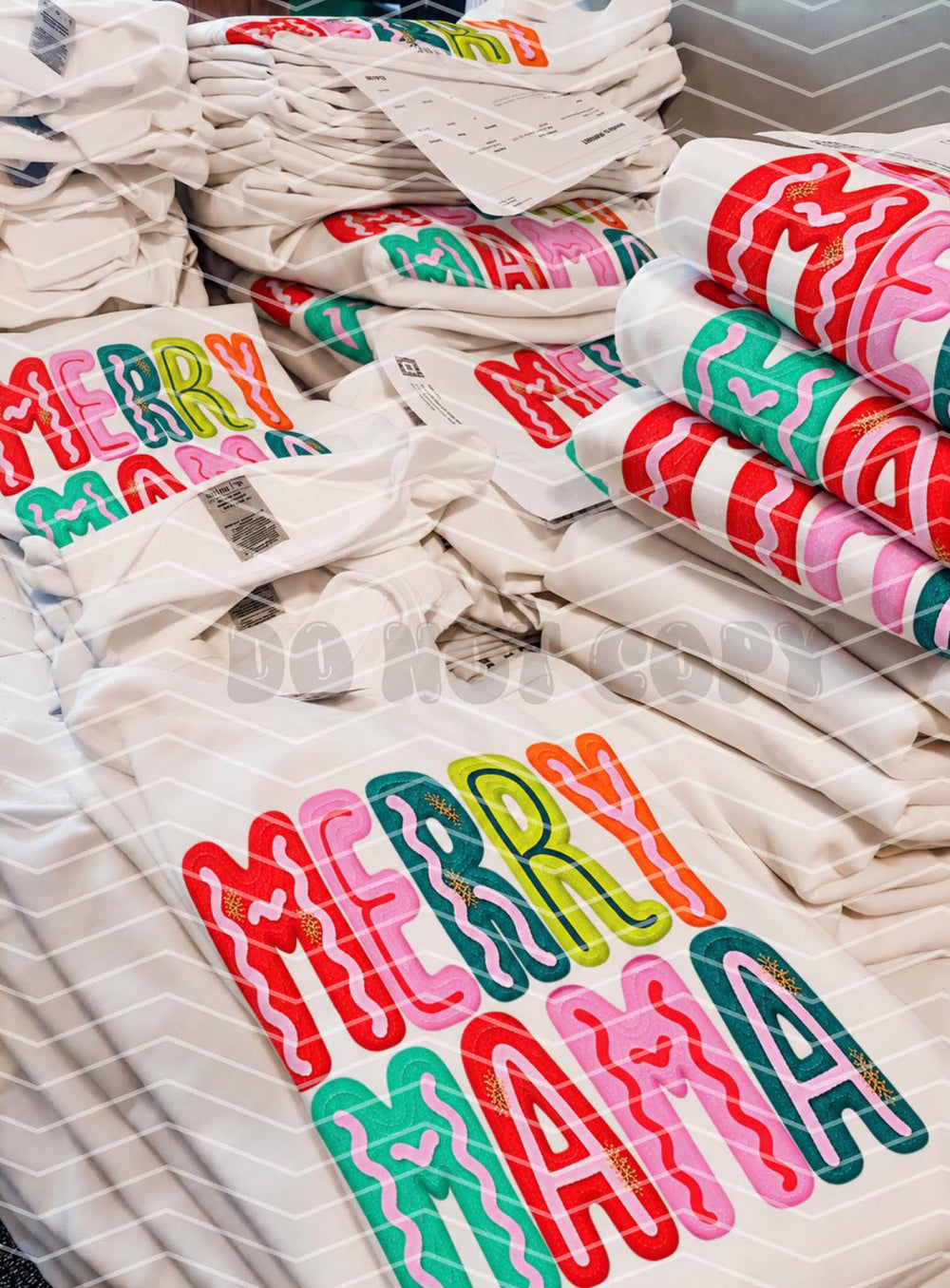 Stacks of white tees and sweatshirts printed with the bright Merry Mama squiggle Christmas design